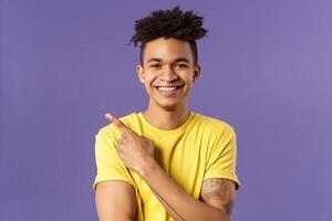 Close-up portrait of enthusiastic, happy young hipster male with dreads, beaming smile and pointing finger upper left corner, present cool product, introduce something really good, purple background photo
