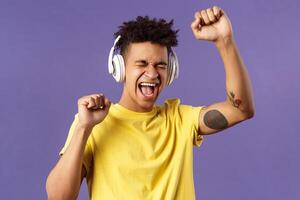 Close-up portrait of cheerful, happy young dancing guy lift hand up singing along, close eyes and smiling upbeat as listening awesome song in headphones, enjoying music, purple background photo