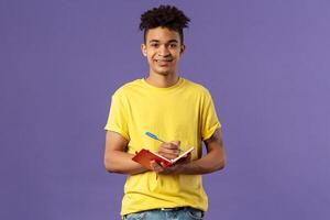 Portrait of young hispanic male student studying online courses, writing down lecture, making personal schedule or taking notes in planner, look camera enthusiastic and interested, purple background. photo