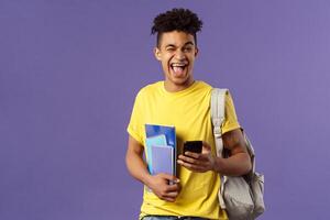 Back to school, university concept. Portrait of joyful, smiling happy man, student using mobile phone, wink at camera upbeat, holding backpack and notebooks, notes from lecture and smartphone photo