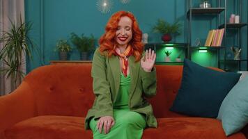 Redhead girl smiling friendly at camera, waving hands gesturing hello, hi, greeting at home on couch video