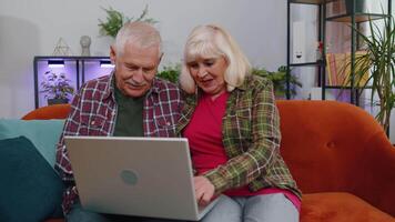 Senior grandmother grandfather using laptop computer, typing at home room, looking at camera smiling video