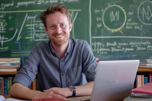 Smiling teacher sits at a desk with a laptop in front of a chalkboard filled with notes photo
