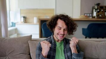 Happy brunette guy in a checkered shirt with curly hair rejoices at his victory while working using a laptop on the sofa in a modern apartment video