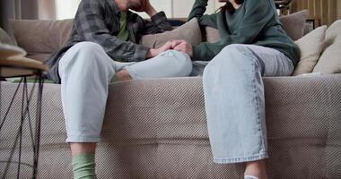 Zoom In bottom view of a happy guy brought with curly hair holding hands with his girlfriend and actively laughing while talking while sitting on the couch at home video
