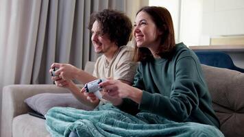 Happy brunette girl together with her boyfriend with curly hair wrapped with a blanket and playing games while sitting on the sofa using a joystick at home video