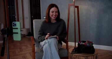Confident brunette girl communicates with her viewers while blogging and sits on a chair and takes notes using a notepad in a modern cozy apartment video