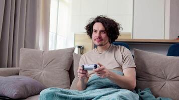 Happy brunette guy with curly hair and mustache plays games using a white joystick while sitting on a sofa with a blue blanket in a modern apartment video