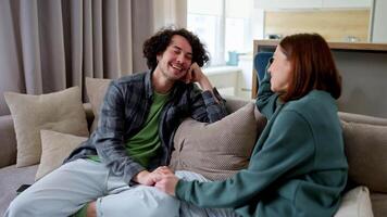 Happy guy with curly hair laughs loudly and has fun while communicating with his brunette girlfriend while sitting on the sofa at home in a modern apartment video