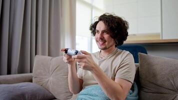 Happy brunette guy with curly hair and mustache plays games and wins with a joystick and sits on the sofa at home in a modern apartment video