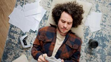 un confidente morena chico con Rizado pelo en un a cuadros camisa hace notas en su cuaderno y luego lágrimas fuera un sábana desde eso y lo hace eso de nuevo mientras acostado en el piso en un almohada alrededor hojas de video