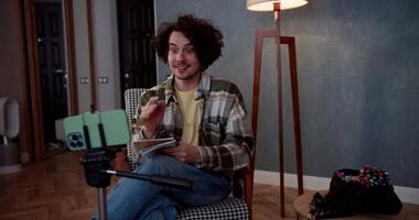 A happy brunette guy with curly hair watches videos and blogs using a smartphone and makes notes in his notebook while sitting on the sofa at home in a modern apartment