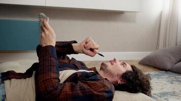 A thoughtful guy comes with curly hair lying on a pillow and writing down his ideas in a notepad while lying on the floor in a modern apartment video