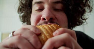 Close-up portrait of a Happy guy with curly hair and mustache biting a burger and enjoying it in the morning in a modern apartment video