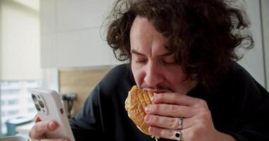 Zoom in confident brunette guy with curly hair and mustache sitting and eating a sandwich while browsing social networks at home during breakfast in the kitchen video