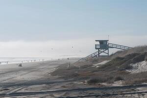 A lone lifeguard tower stands on a sandy beach, overlooking a misty ocean. A few people are scattered along the shore, enjoying a sunny day. photo
