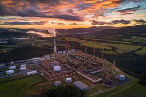 An oil refinery nestled in a valley, illuminated by the setting sun, with smoke rising from a tall stack. photo