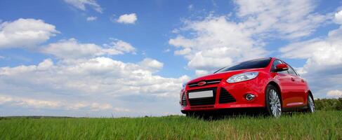 Red car on a blue sky background with white clouds. The car is standing in the field on the green grass. photo