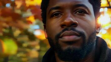 An African American man with a bold beard and a serious expression stands against a tree trunk covered in autumn leaves the rich colors providing a striking contrast.. video
