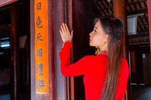 Vietnamese girl with Ao Dai dress standing in traditional house of Vietnam. Text in photo mean best wishes to family, happiness, prosperity, health. Tet holiday and New Year.
