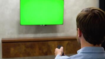 Young Man is Sitting on a Sofa, Choosing Channel and Watching TV with Horizontal Green Screen Mock Up. View From The Back video