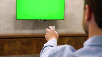Young Man is Sitting on a Sofa, Choosing Channel and Watching TV with Horizontal Green Screen Mock Up. View From The Back video