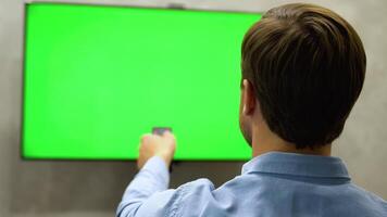 Young Man is Sitting on a Sofa, Choosing Channel and Watching TV with Horizontal Green Screen Mock Up. View From The Back video