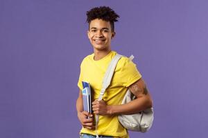 Back to school, university concept. Portrait of handsome charismatic hispanic guy, student walking to campus, going for lecture, studying hold backpack and notebooks, stand purple background photo