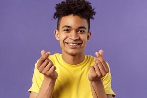 Close-up portrait of lovely cute young man with afro haircut, showing korean heart finger signs and smiling, express his love and sympathy towards person, stand purple background photo