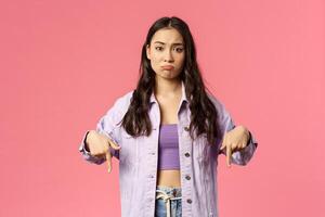 Displeased skeptical young asian girl pointing fingers down, pounting and making disappointed face, missed chance, lose in competition or see bad result, standing pink background unimpressed photo