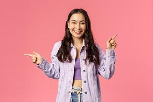 Portrait of stylish assertive cute korean girl giving choice, pointing fingers left and up, promote products, give advice where find useful links on online shops, internet stores, pink background photo