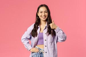 I am in. Cheerful confident pretty, stylish asian girl supports you, show thumb-up and smiling with approval, nod agree friend made good point, like and accept plan, pink background photo