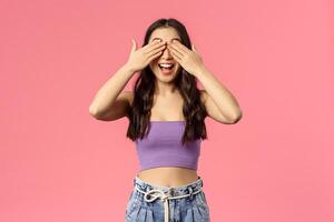 Portrait of excited enthusiastic good-looking woman in crop-top, awaiting for command to open eyes, smiling amused, blindfolded waiting surprise gift, standing pink background photo
