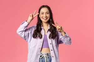 Kawaii asian girl sending good positive vibes, enjoying bright sunny weekend, having fun, partying and brighten up your day with beaming happy smile show peace gesture, pink background photo