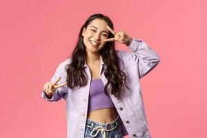 Portrait of charismatic good-looking asian girl sending positive vibes, show peace kawaii gesture near face and dancing, smiling inviting people to visit her store or apply company, pink background photo