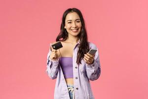 Portrait of young stylish girl, smiling pleased camera, using mobile phone and hold credit card to pay for online purchase, shop in internet, order food delivery, standing pink background photo