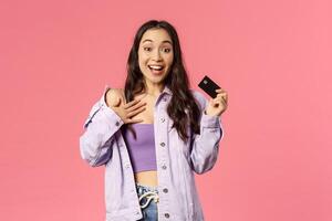 Portrait of excited, enthusiastic young girl holding credit card, gasping and touch chest as being overwhelmed with new clothes collection at online store, ready to buy and waste money photo