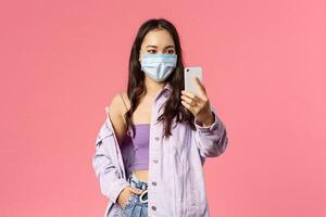 Covid19, quarantine, people concept. Stylish young woman staying safe, wear face mask to go outside grocery shopping, taking selfie on mobile phone, talking with siblings online during pandemia photo