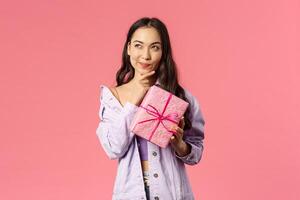 Portrait of happy thoughtful young asian girl receive wrapped box with gift, wonder what hidden inside, trying to guess, smile and look up thinking, standing pink background intrigued photo