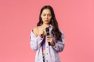 Portrait of awkward silly asian girl tap microphone as checking is it working, her turn to sing song on karaoke party, frowning complicated, standing pink background unsure how this thing works photo