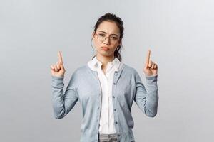 Skeptical, disappointed cute asian girl dont recommend this product, pointing fingers up with bothered, complaining and gloomy expression, have negative thoughts about company, grey background photo