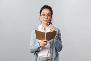 Education, teachers, university and schools concept. Thoughtful smart asian female student studying, prepare homework or write down lecture in notebook, holding planner, thinking over exercise photo