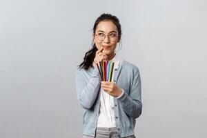 Hobbies, creativity and art concept. Scheming, creative and smart young talented student thinking-up what draw next, holding colored pencils, smirk and look up thoughtful, grey background photo