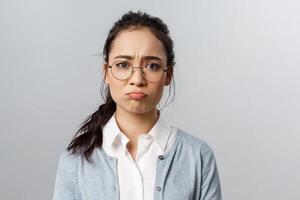 Close-up portrait of gloomy, upset and disappointed young asian female facing failure, losing, regret mjssing interesting event, pouting and sob, frowning camera standing grey background photo