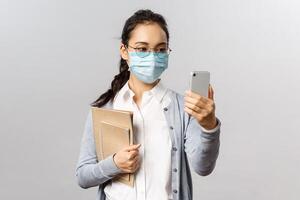 Covid19, virus, health and medicine concept. Young asian girl in medical mask, holding notebooks and work reports, taking selfie on smartphone, videocalling friends while social distancing photo