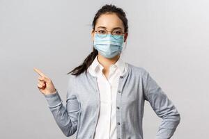 Covid19, virus, health and medicine concept. Portrait of young serious-looking asian girl in medical mask, prevent spreading coronavirus and influenza, pointing left, showing info about disease photo