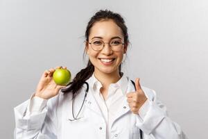 Portrait of young cheerful asian female doctor, therapist or diatologist talking abut healthy diet, showing green apple and thumb-up, recommend eat fruits, smiling encourage look after health photo