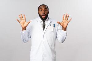Portrait of shocked and nervous african-american male doctor raising hands in surrender, trying calm down tensed family of patient, look insecure and unaware what do, stand grey background photo