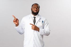 Covid19, healthcare and clinic concept. Portrait of confused and displeased, judgemental african-american doctor, grimacing in aversion, cringe looking and pointing left, patient not wearing mask photo