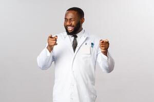 Portrait of cheeky smiling, handsome african-american male doctor flirting or being cheeky, winking pointing at camera with pleased grin, talking about you, inviting for check-up, ecnourage vaccinate photo